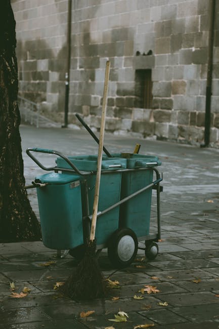 A commercial cleaning cart positioned on a paved outdoor area next to a tree, containing two large teal plastic bins with attached handles, surrounded by cleaning tools such as brooms and mops. The ca