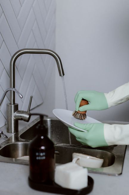 Close-up of a person wearing yellow rubber gloves using a blue sponge to clean a white wall or tiled surface in a kitchen, with a spray bottle of cleaning solution held in their other hand. The surfac