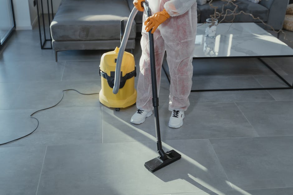 A professional cleaner wearing white protective clothing and orange gloves is performing surface cleaning on a large, grey tile floor in a modern living room. They are using a vacuum cleaner with a ye